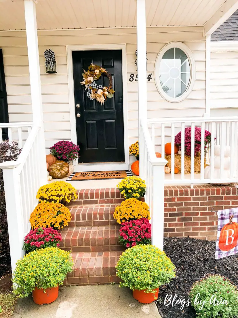 fall front porch decorated with an assortment of mums lining the steps to a front door decorated with a fall wreath complete with fall yard flag